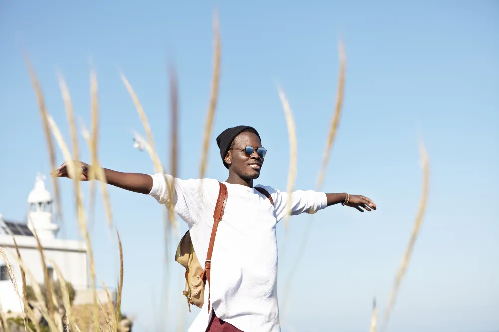 Cheerful stylish young Afro American tourist with knapsack wearing hat and stylish sunglasses